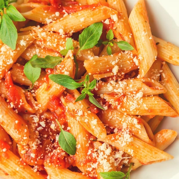 Penne pasta with red tomato sauce, grated cheese, and fresh basil leaves in a white bowl.